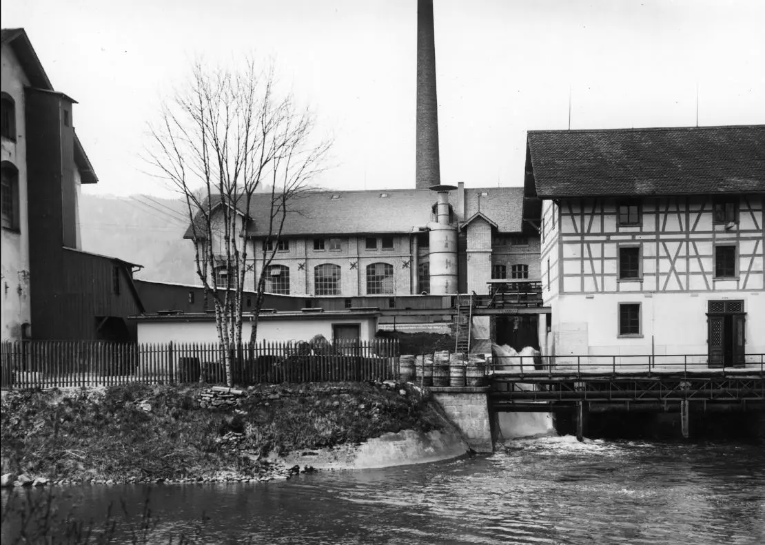 Fotografia storica in bianco e nero della cartiera sulla Sihl: edifici della fabbrica e un alto camino dietro uno sbarramento sul fiume